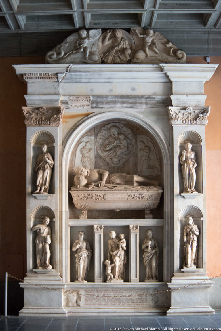 White stone wall tomb with recumbent effigy, arched recess, relief carving, and small figures in niches.