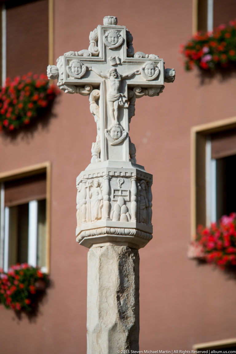 Carved pale stone cross with relief figures on a decorated pedestal against a warm building façade.