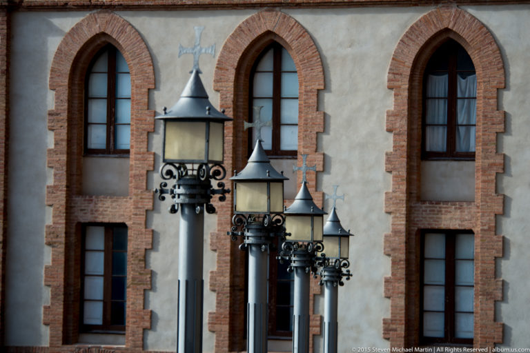 Four decorative black metal lamps stand before a pale wall with tall brick-framed pointed windows.