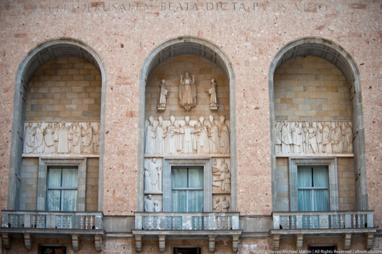 Three arched façade bays with pale religious relief sculpture, windows, balconies, and a Latin inscription above.