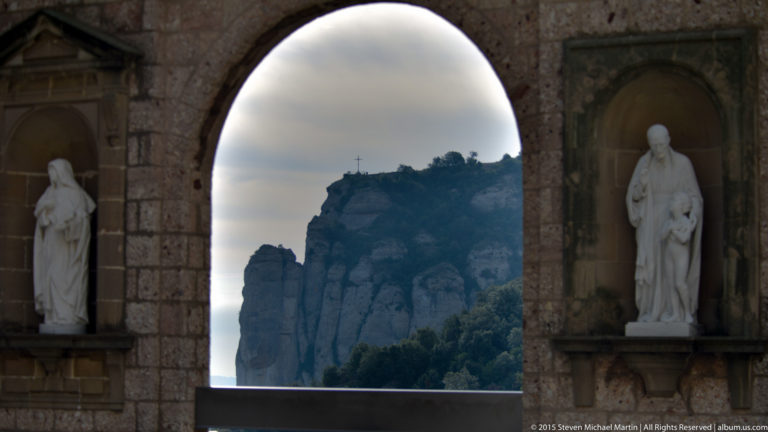 Distant cross on a Montserrat summit framed by a tall arch, with saint statues in side niches.