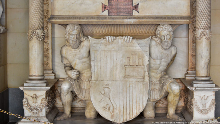 Carved stone coat of arms flanked by two muscular male figures within an ornate architectural surround.