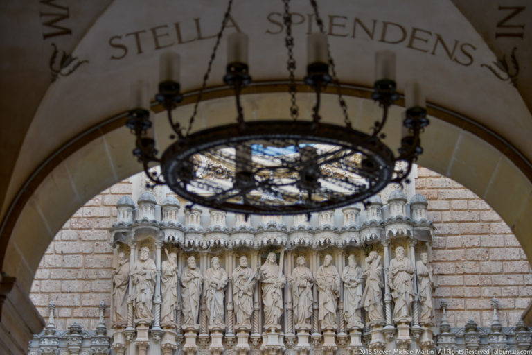 Christ and Apostle statues on Montserrat basilica façade seen through an arch beneath a circular hanging fixture.