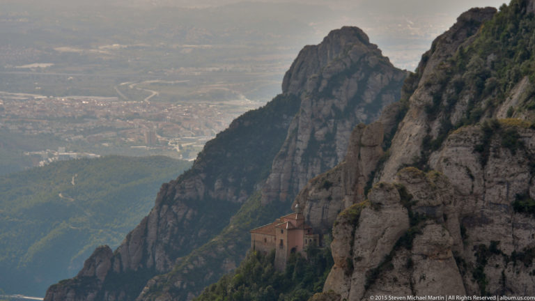 Small hermitage building clings to a narrow ledge among steep Montserrat cliffs above a distant valley.