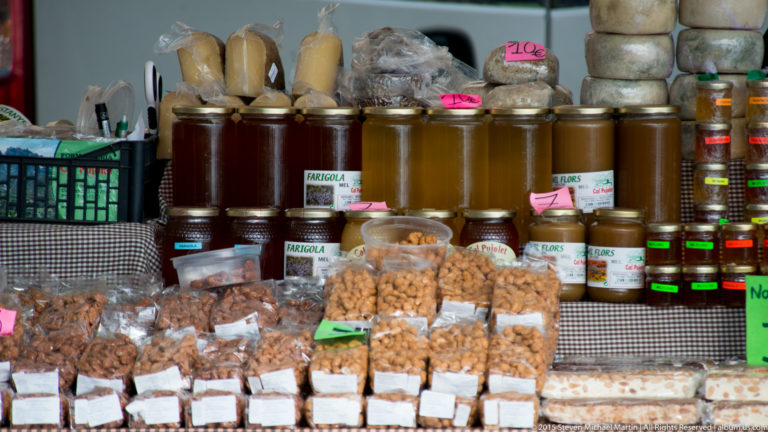 Market stall displaying jars of honey, bags of nuts, and packaged confections with handwritten labels and prices.