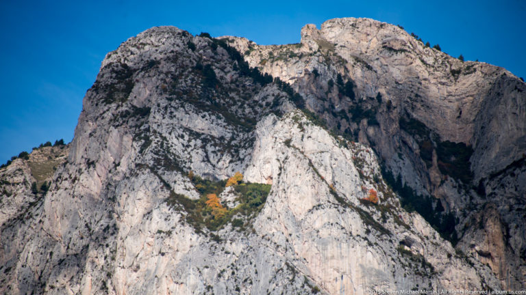 Massive pale fractured cliffs of Montserrat fill the frame, with sparse vegetation and small autumn-colored trees on ledges.