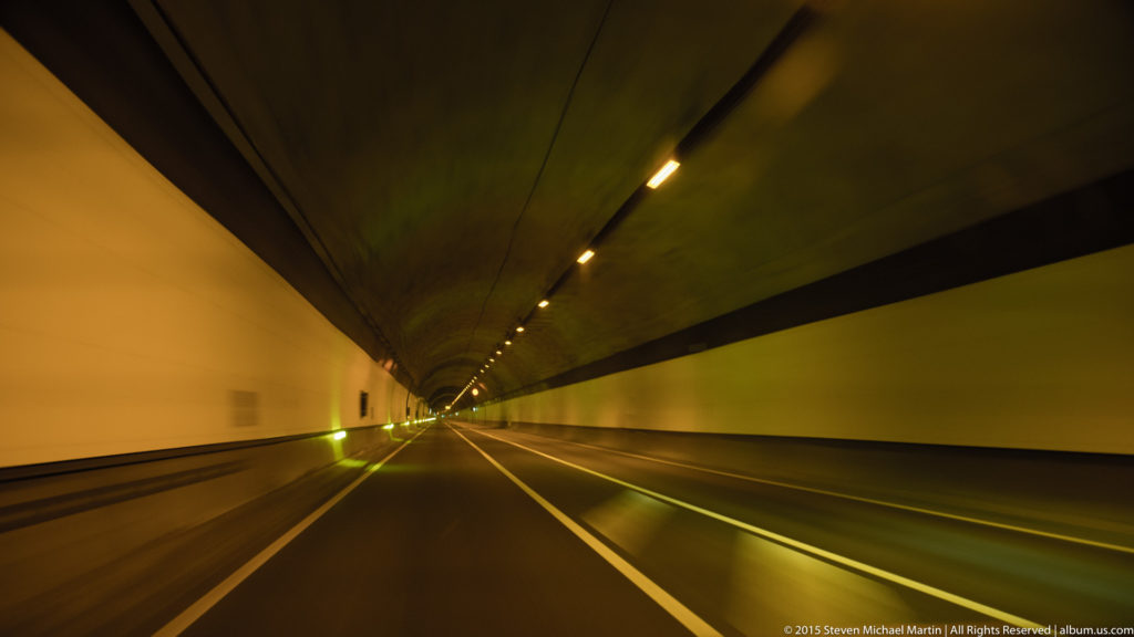 Long illuminated road tunnel with converging lane lines and ceiling lights leading toward a distant vanishing point.
