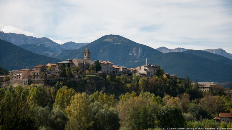 Compact hilltop village with church bell tower stands above woodland against layered mountain ridges.