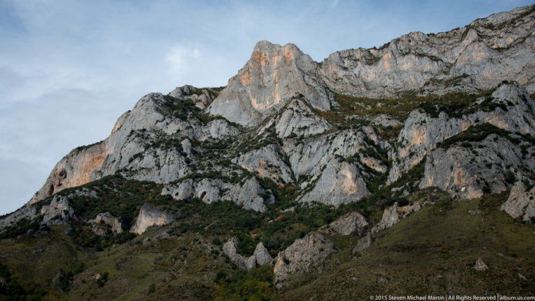 Rugged pale rocky ridge with dark vegetation and an orange-lit outcrop beneath a hazy sky.