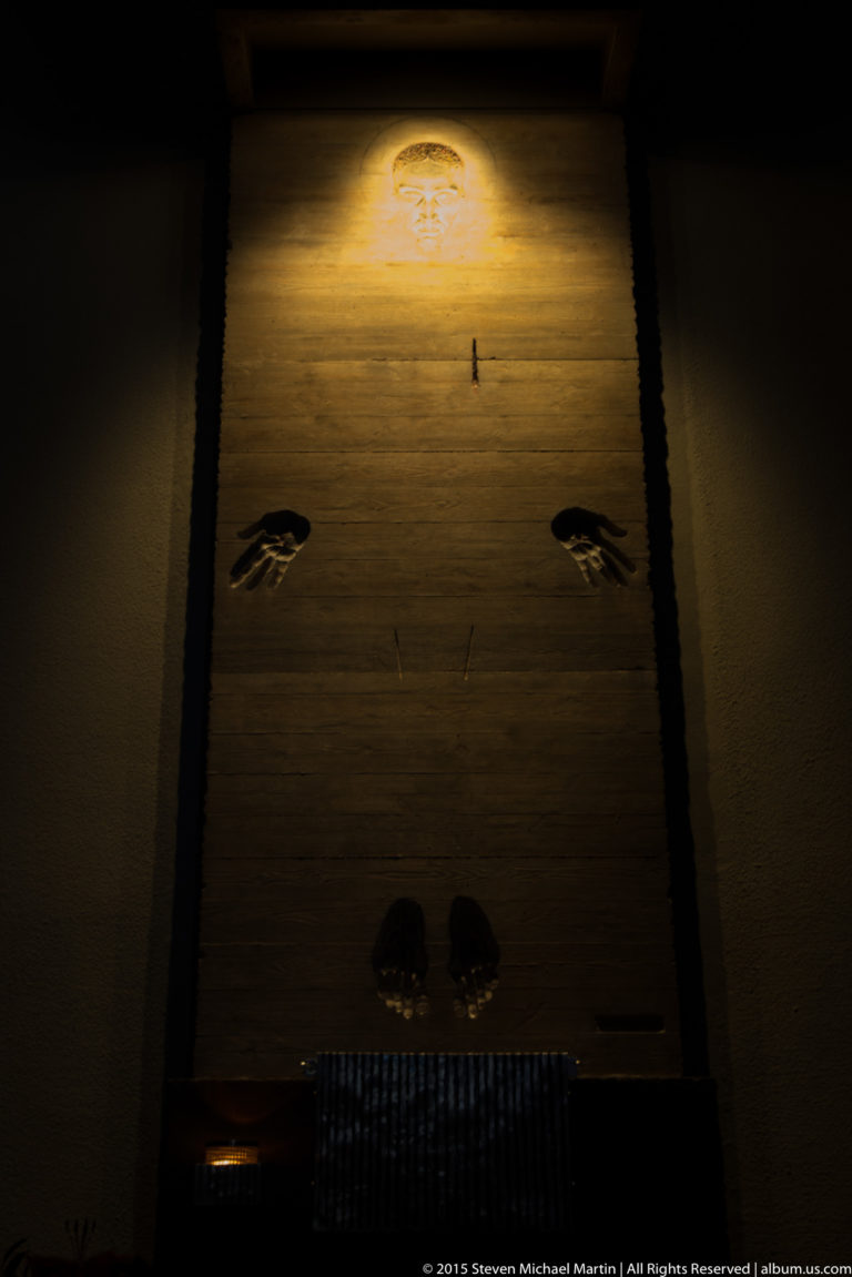 Dark interior wall lit from above, with paired hand motifs, a small central cross-like element, and lower dark forms.