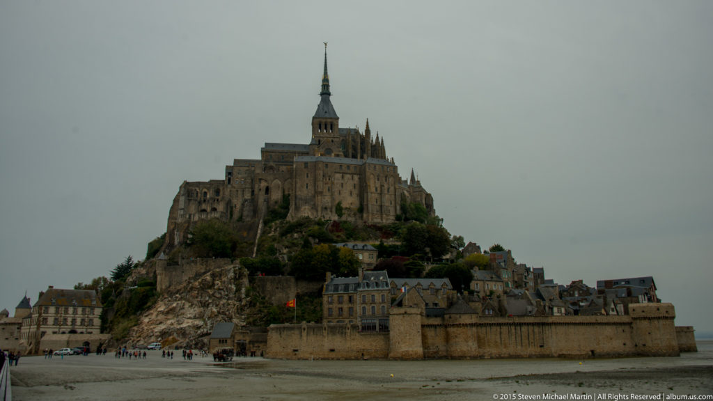 Mont Saint-Michel rising above its walled village and tidal flats beneath an overcast sky.