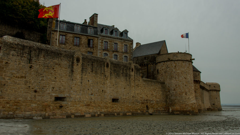 Stone defensive walls and a round tower at Mont Saint-Michel, with buildings rising above them.