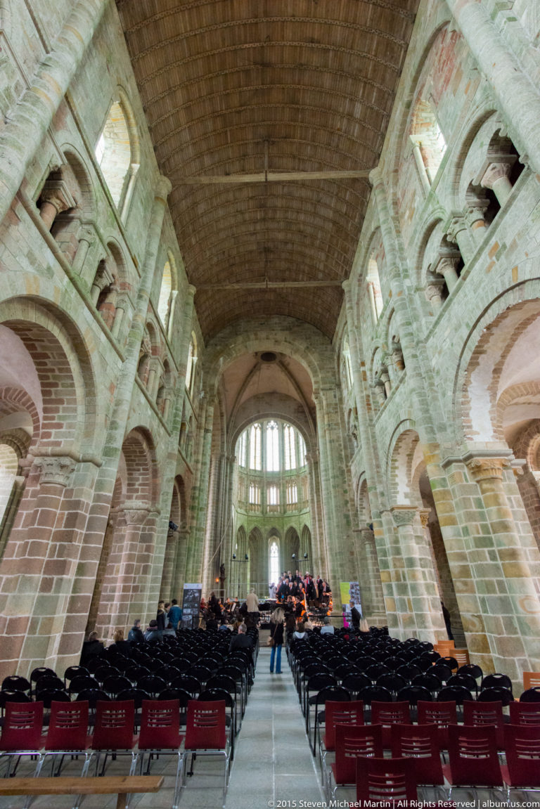 Long nave of the abbey church with stone arcades, timber ceiling, rows of chairs, and distant choir.