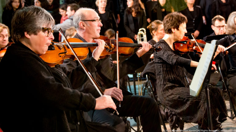 Close view of violinists performing in an orchestra, with bows raised and other musicians behind them.