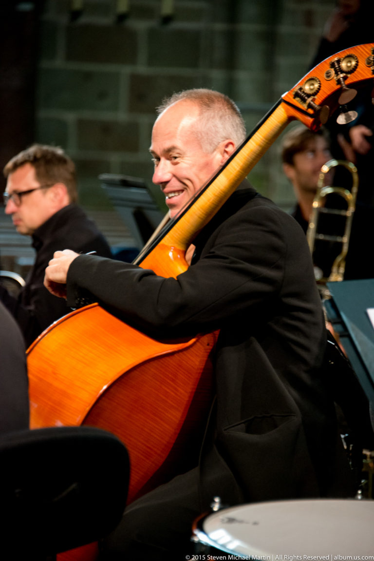 Smiling double bass player performing, with the large wooden instrument angled across the frame.