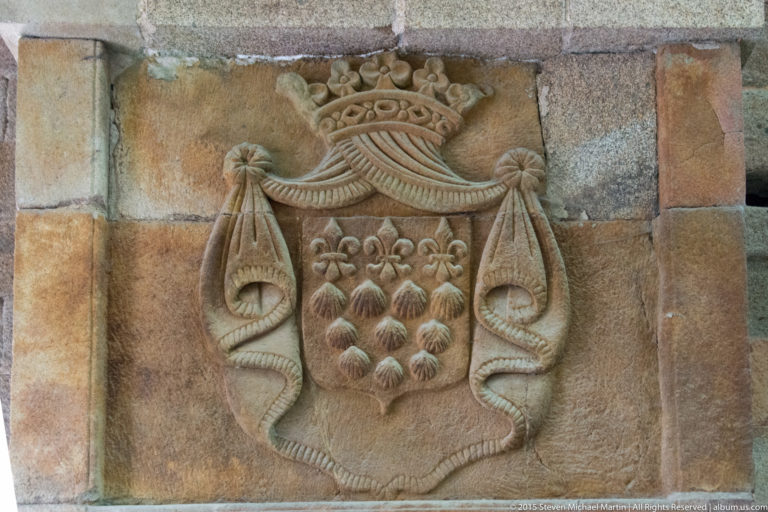 Weathered carved stone coat of arms with a crown, shield, floral forms, and hanging tassels.