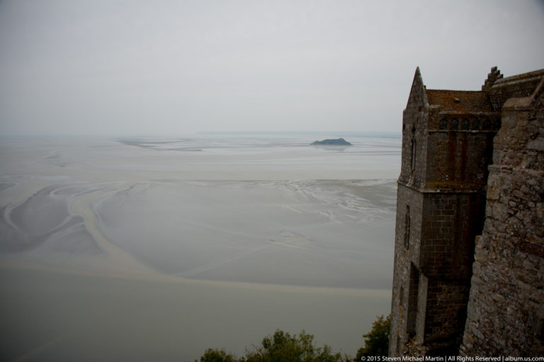 Pale tidal flats and channels of Mont Saint-Michel Bay seen beside the edge of a stone abbey wall.