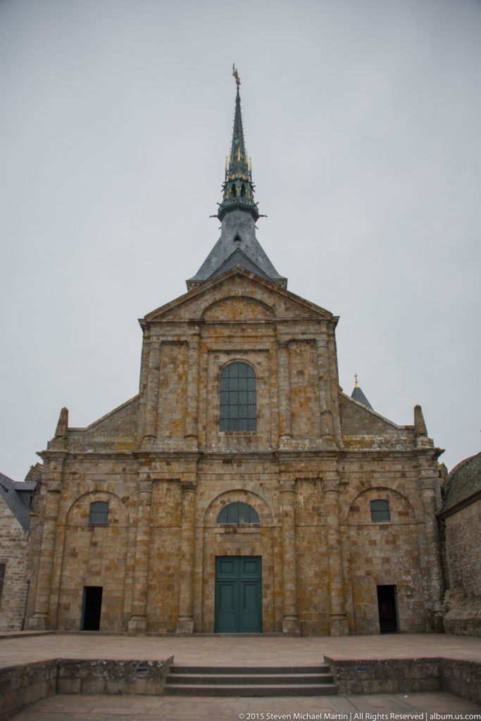 Stone west façade of the abbey church with central doorway, large windows, and slender spire.