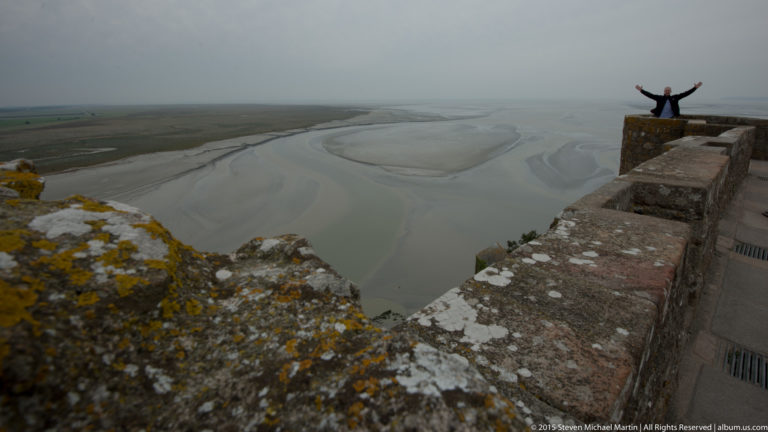 Visitor with raised arms standing on a stone parapet above misty tidal flats and channels.