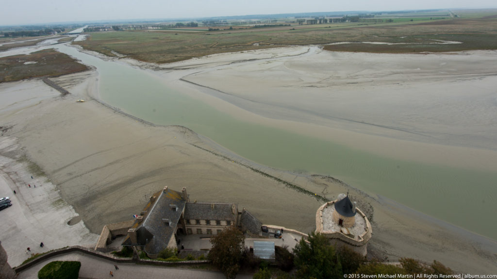 High view of a pale tidal channel, sandy flats, and buildings at the base of Mont Saint-Michel.