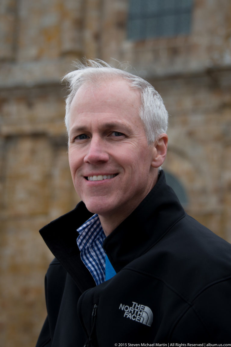 Close portrait of a smiling gray-haired man in a dark jacket, with stone architecture blurred behind.