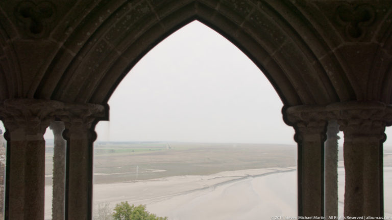 Pointed stone cloister arch framing the pale tidal flats of Mont Saint-Michel Bay.