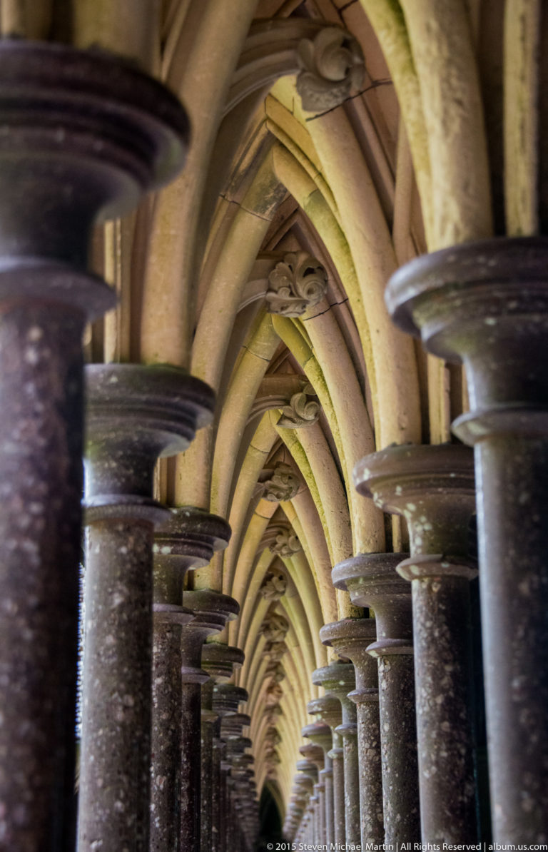 Close oblique view along repeating dark columns and pointed arches in the abbey cloister.