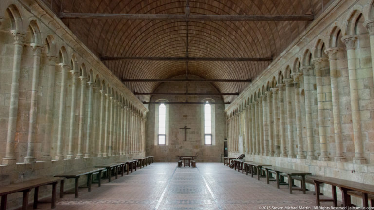 Long monastic refectory with timber ceiling, narrow windows, rows of tables, and stone walls.