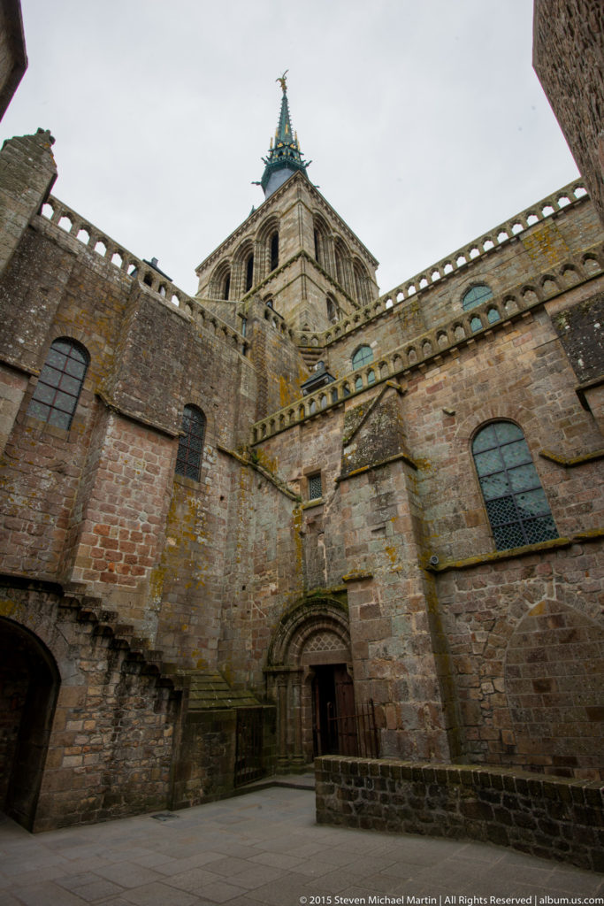 Upward view of massive stone abbey walls, windows, buttresses, and the church tower at Mont Saint-Michel.