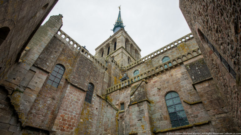 Church tower and spire seen from below between massive stone walls of Mont Saint-Michel Abbey.