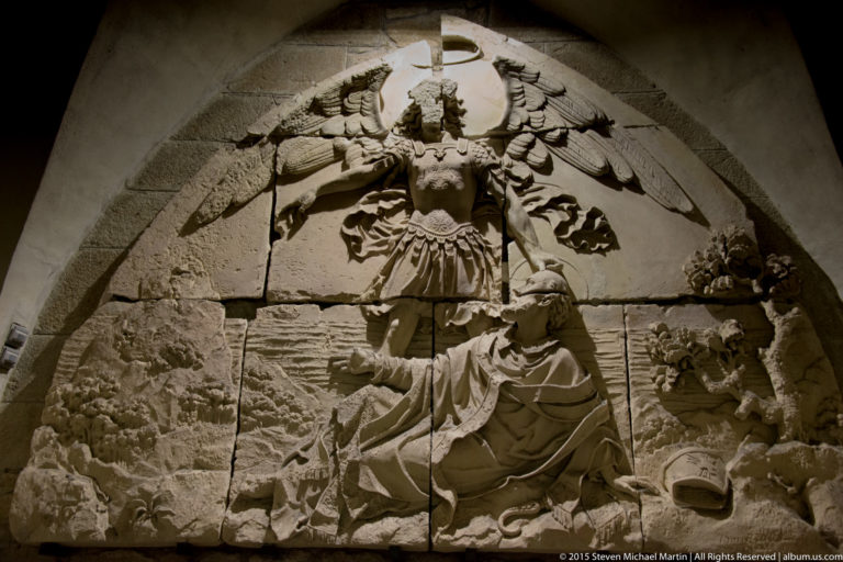 Stone relief of the winged Archangel Saint Michael standing over a defeated dragon-like figure.