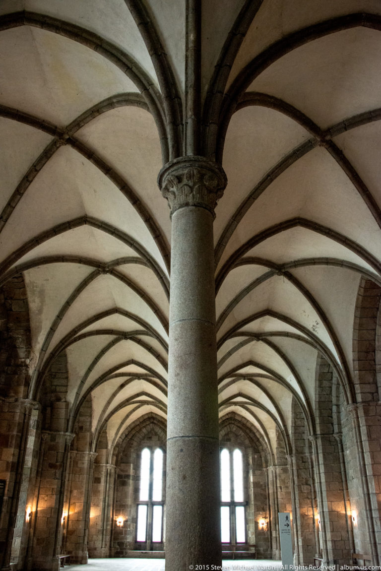 Central stone column supporting low ribbed vaults across a broad hall in Mont Saint-Michel Abbey.