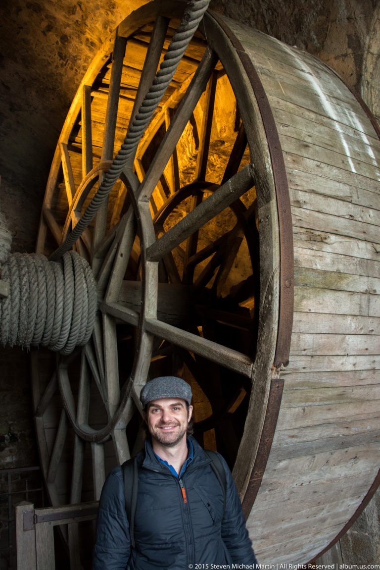 Man in a flat cap standing beside a towering wooden treadwheel and thick rope inside the abbey.