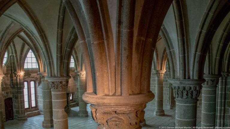 Clustered stone columns, carved capitals, and pointed arches inside a Gothic hall at Mont Saint-Michel Abbey.