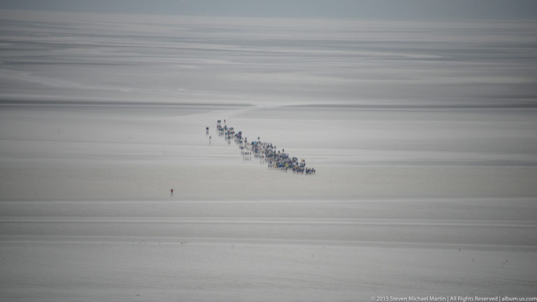 Long distant line of people walking across broad pale tidal flats in Mont Saint-Michel Bay.