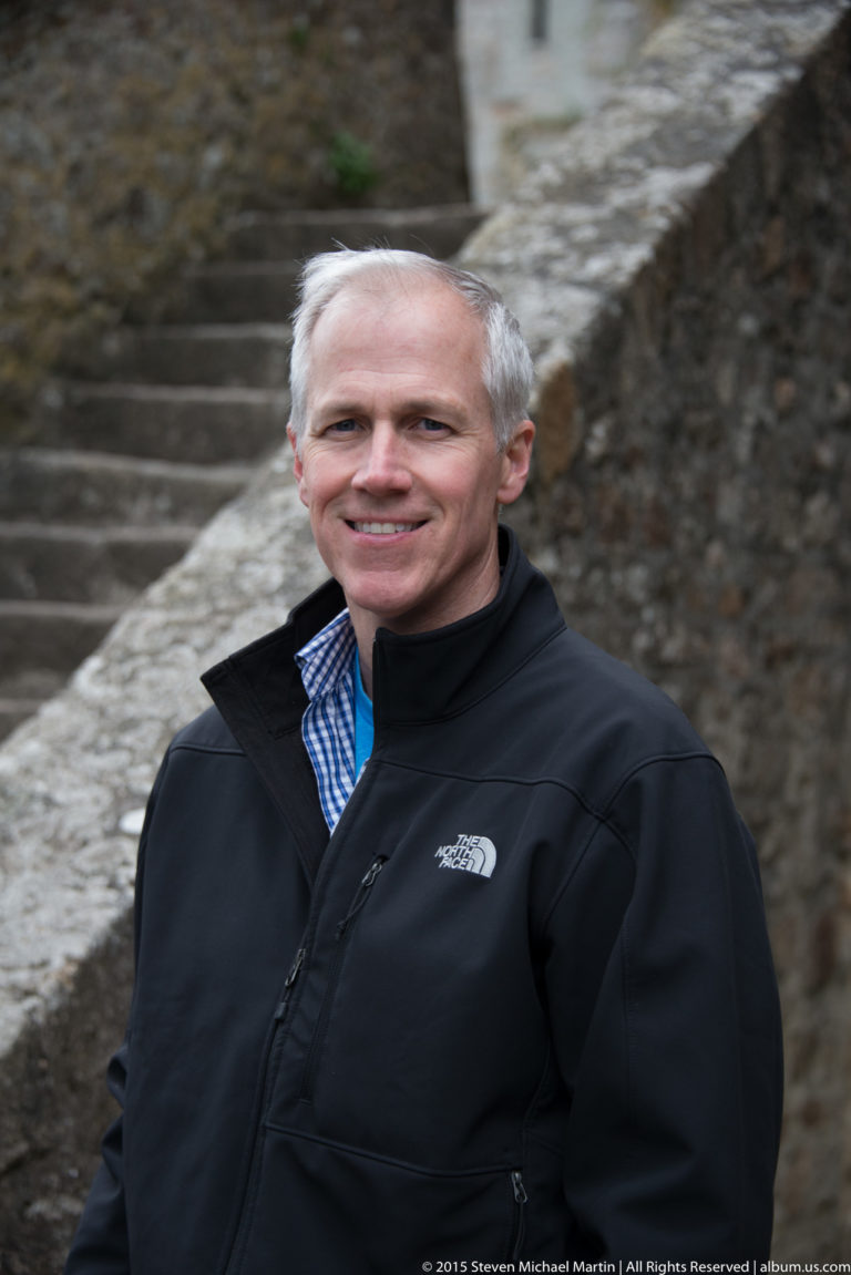 Smiling gray-haired man in a dark jacket standing beside stone steps and walls.