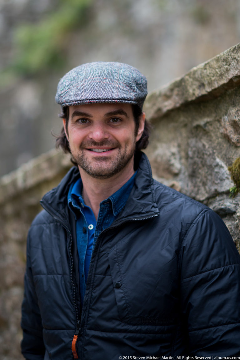 Smiling dark-haired man in a gray flat cap and dark jacket standing before a stone wall.