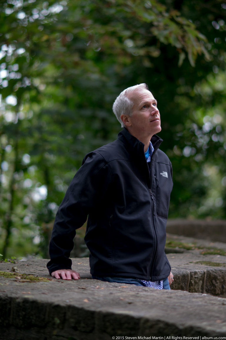 Gray-haired man leaning on a low stone wall beneath green foliage and looking upward.