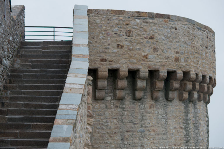 Round stone defensive tower with projecting corbels beside a steep stairway.