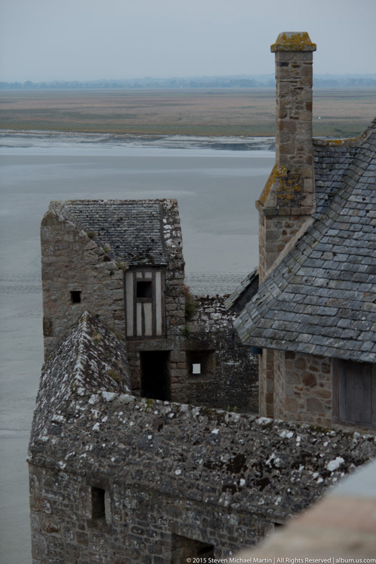 Stone and slate rooftops with chimneys inside Mont Saint-Michel, overlooking broad tidal flats.