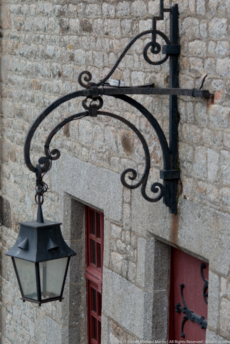 Black hanging lantern suspended from a decorative wrought-iron bracket on a stone wall.