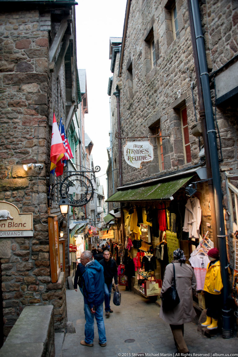 Busy narrow stone street lined with shops, signs, flags, merchandise, and visitors.
