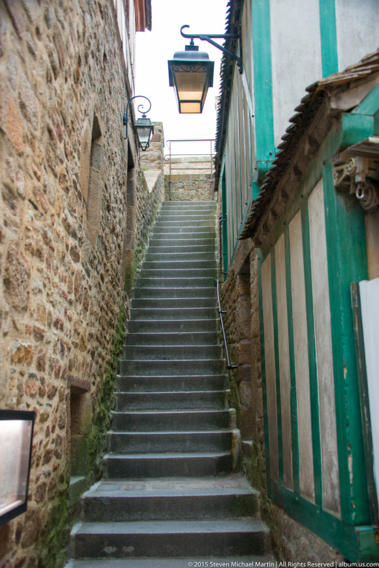 Steep stone staircase rising between a rough stone wall and green-painted half-timbered building.