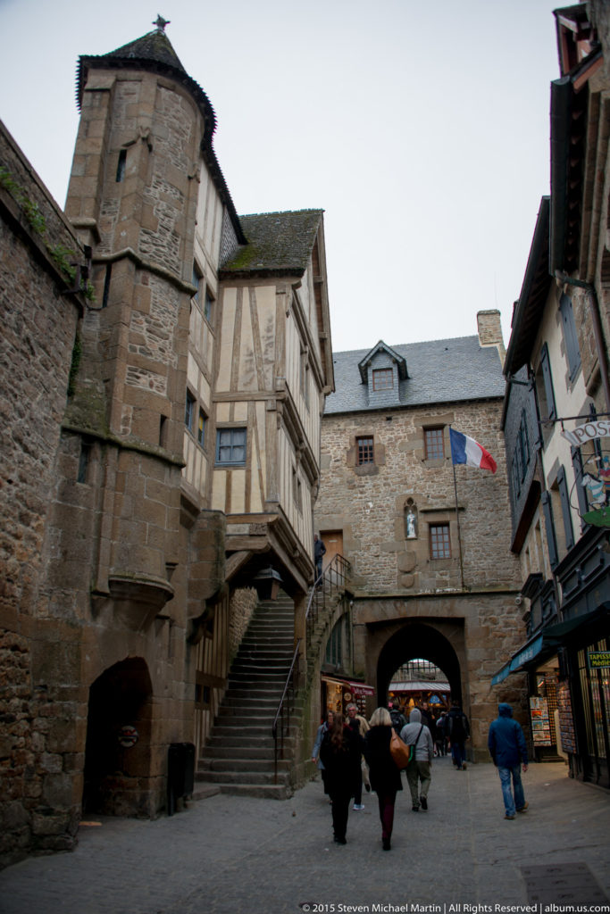 Stone and half-timbered village buildings around steps and an arched passage, with visitors and a French flag.