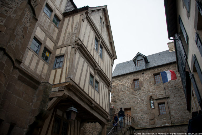 Upward view of a projecting half-timbered house beside stone buildings and a French flag.