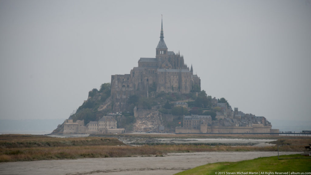 Distant misty view of Mont Saint-Michel rising above flat tidal fields and coastal ground.
