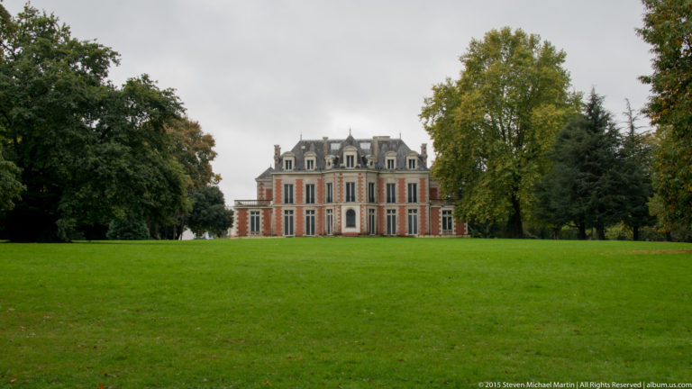 Symmetrical red-brick château with pale stone trim and a slate roof stands beyond a broad green lawn and mature trees.