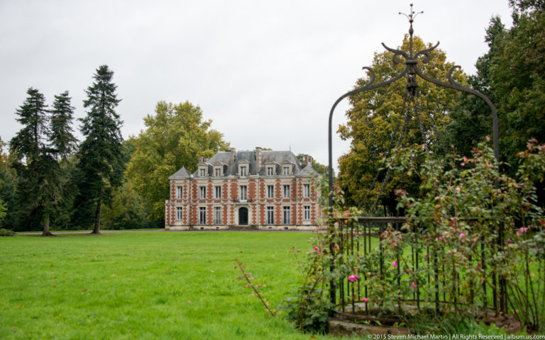 Historic château seen across a green lawn with flower plantings, garden metalwork, and tall trees framing the view.