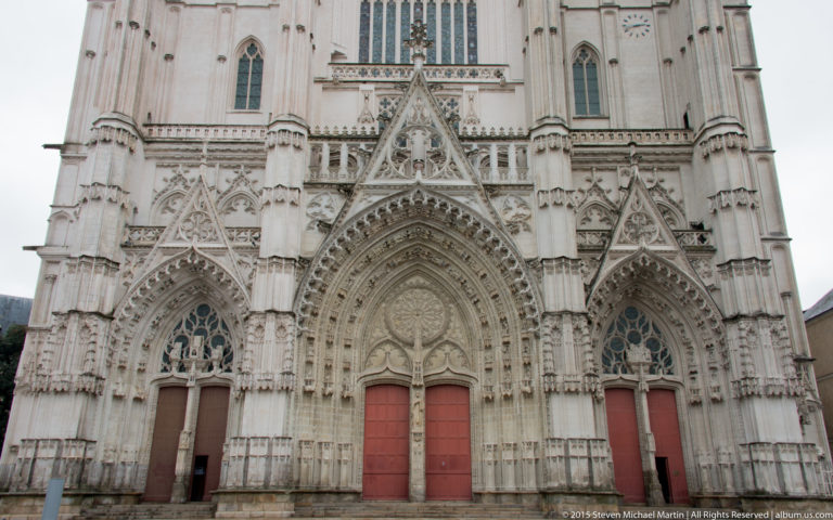 Three richly carved Gothic portals with red doors on the west façade of Nantes Cathedral.