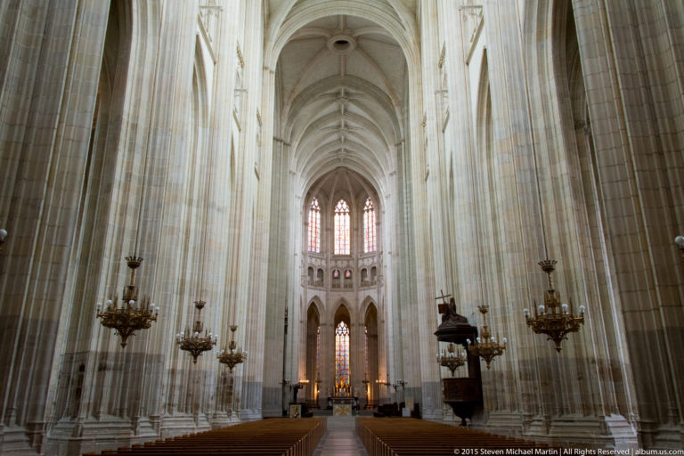 Long central nave of Nantes Cathedral with tall pale piers, ribbed vaults, chandeliers, pews, and stained glass ahead.